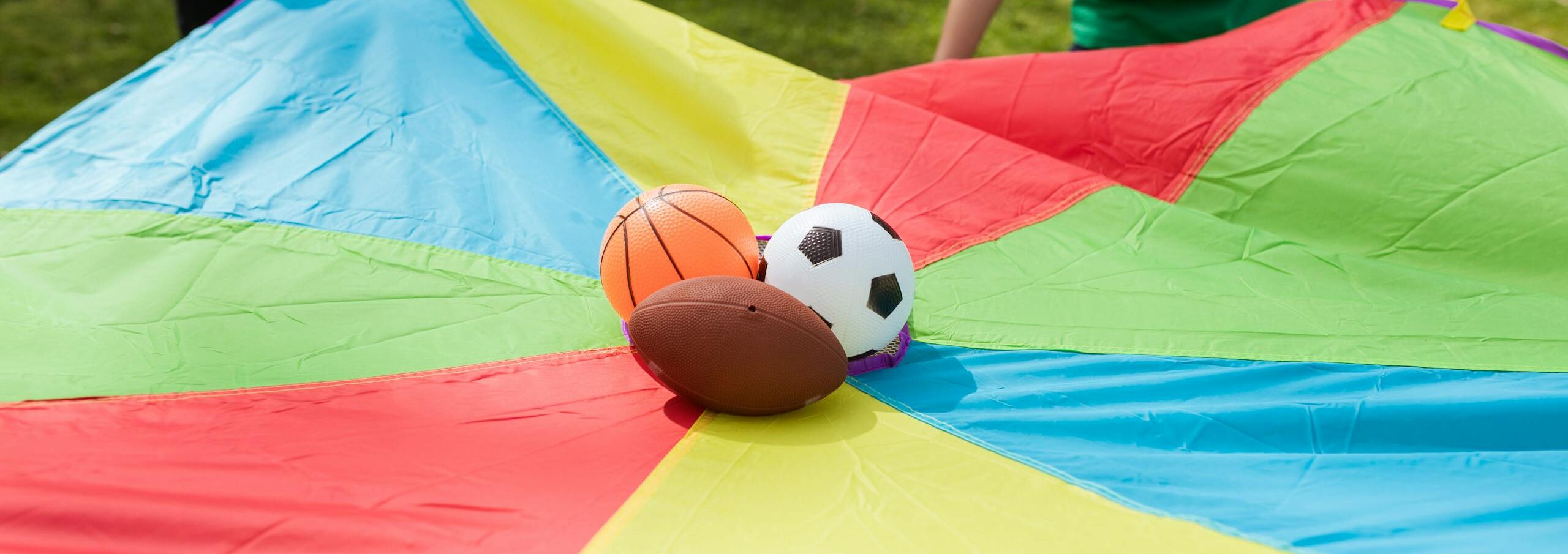 Children playing outside with a colorful parachute and sports balls on grass.
