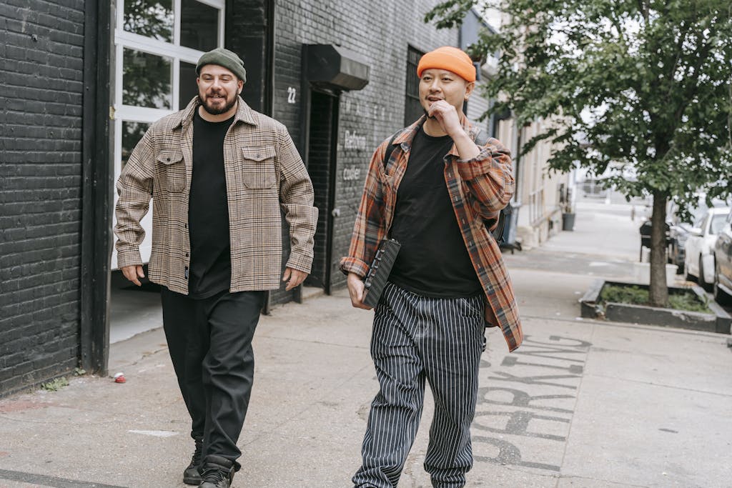 Two men in beanies and plaid shirts walking on a city sidewalk, showcasing urban fashion.
